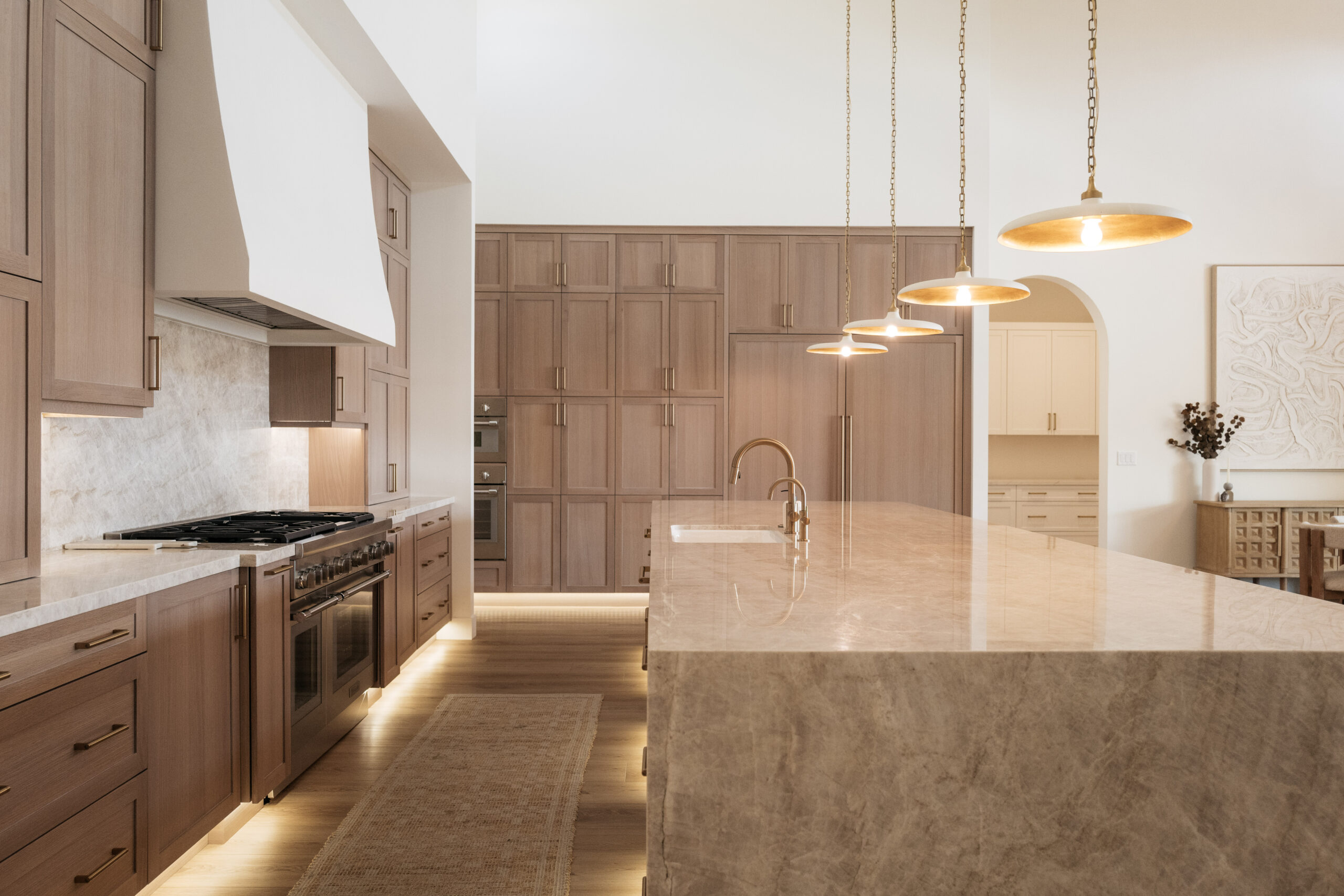 A photo of Pennington Place Kitchen. A large waterfall island of Taj Mahal Quartzite, Rift Sawn white oak cabinetry, white oak plank flooring, limewashed custom range hood, with pendant lights and toe kick lighting.